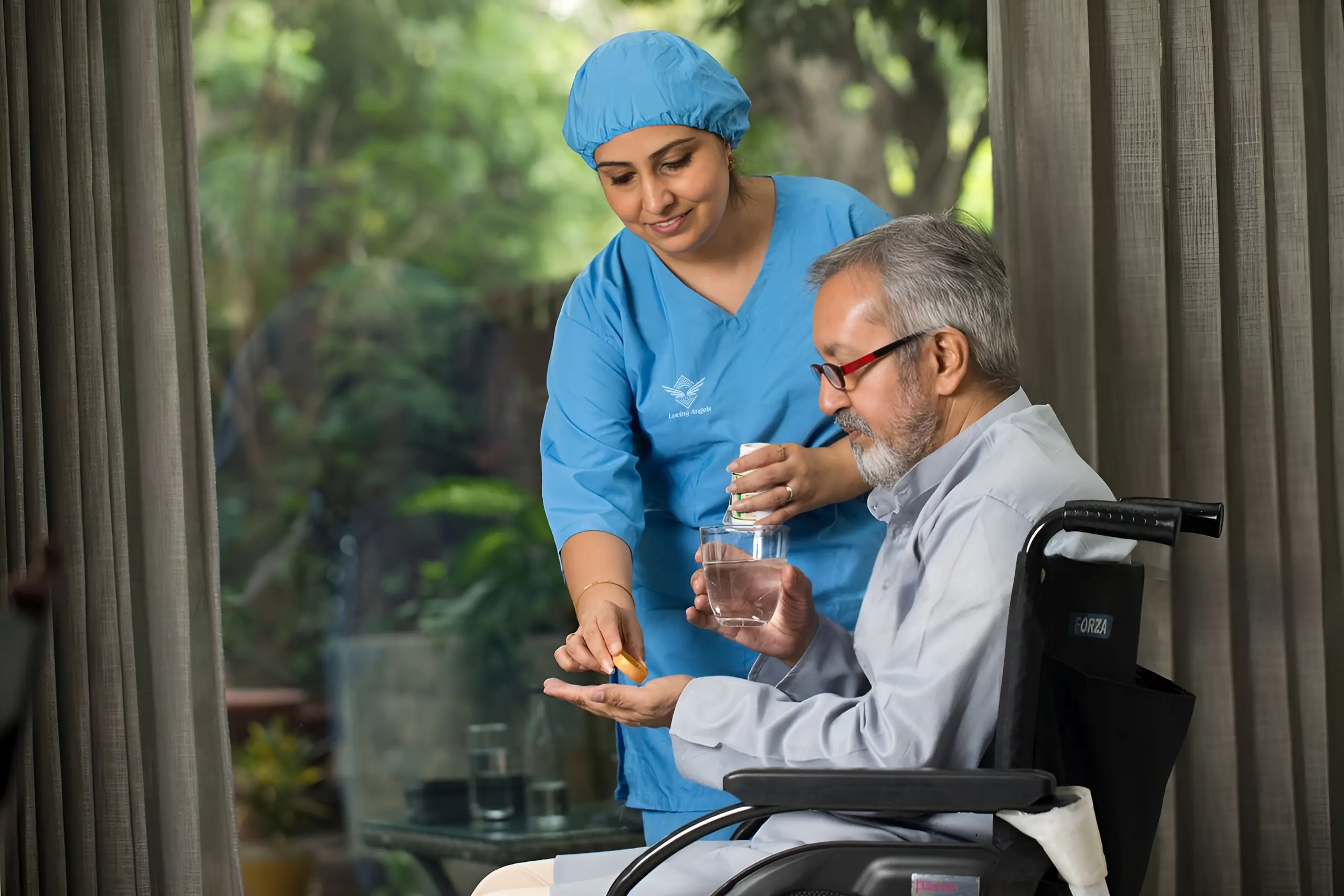 Caregiver in blue scrubs helps elderly man in wheelchair take medication with water.