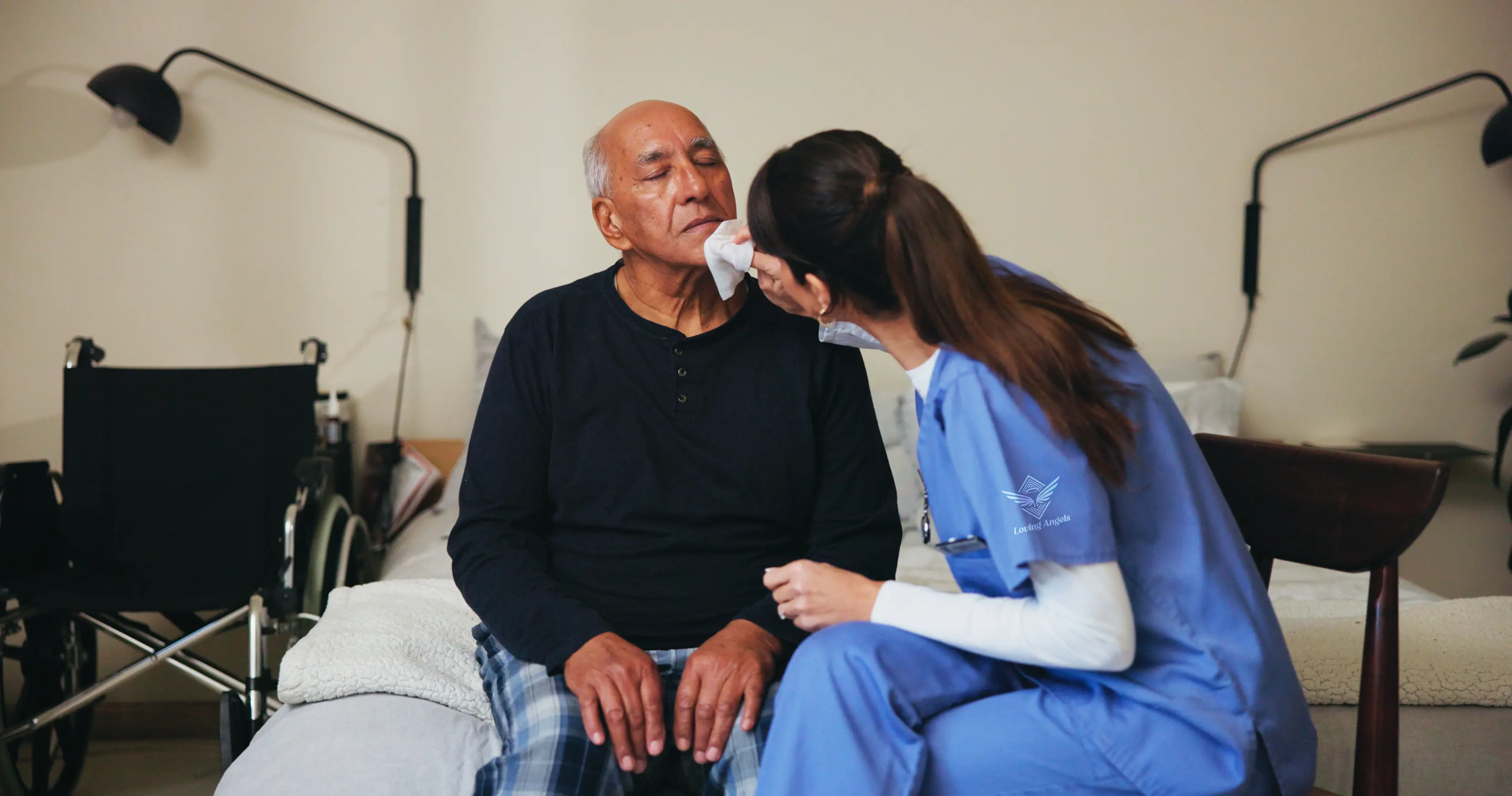 Caregiver in blue scrubs cleaning elderly man's face sitting on a bed with a wheelchair nearby.