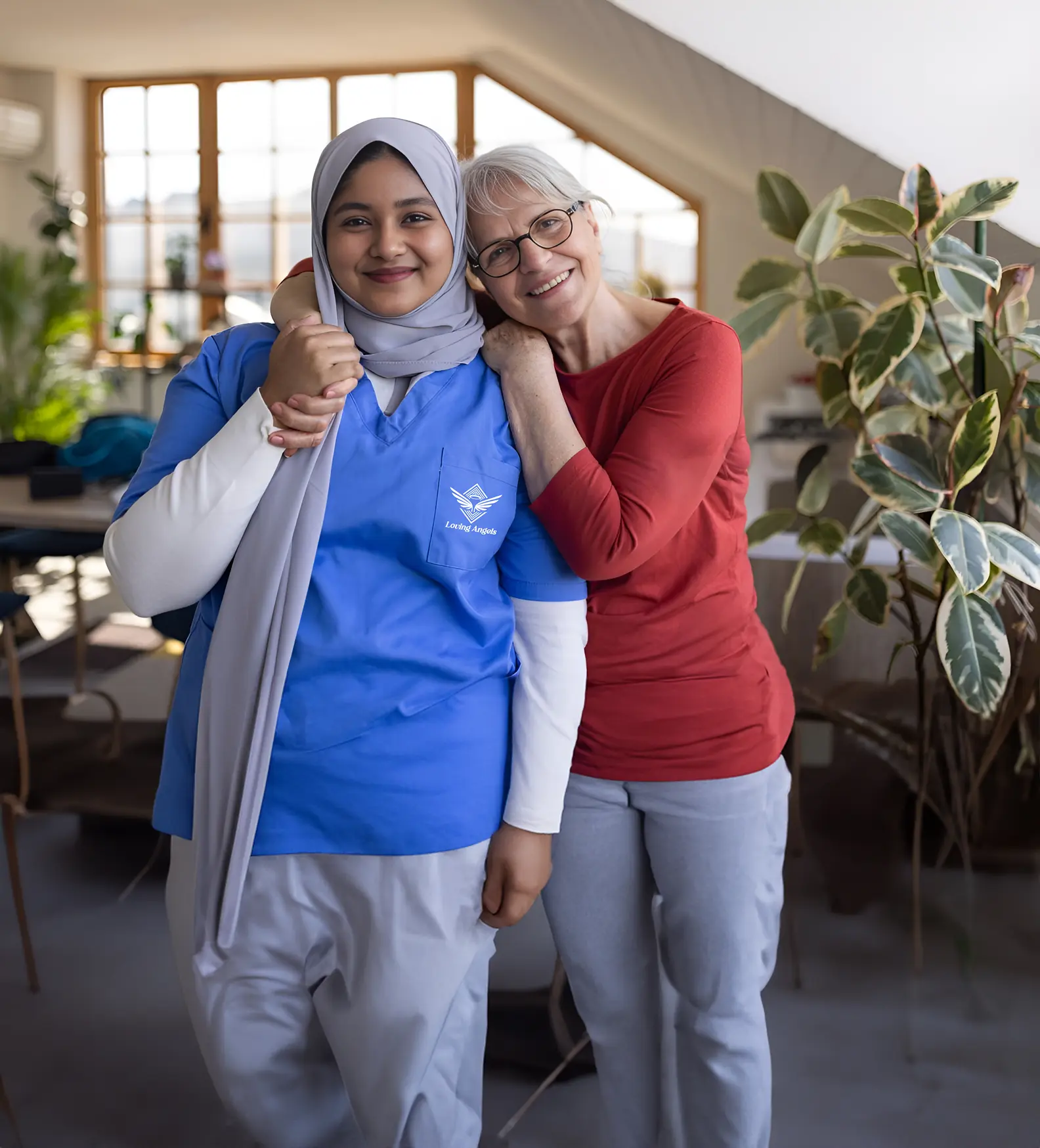 Smiling elderly woman hugging young female caregiver wearing blue scrubs and hijab indoors.