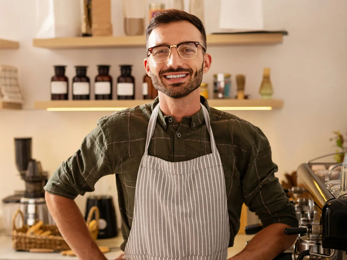 Smiling man wearing glasses and striped apron standing in a coffee shop near equipment.