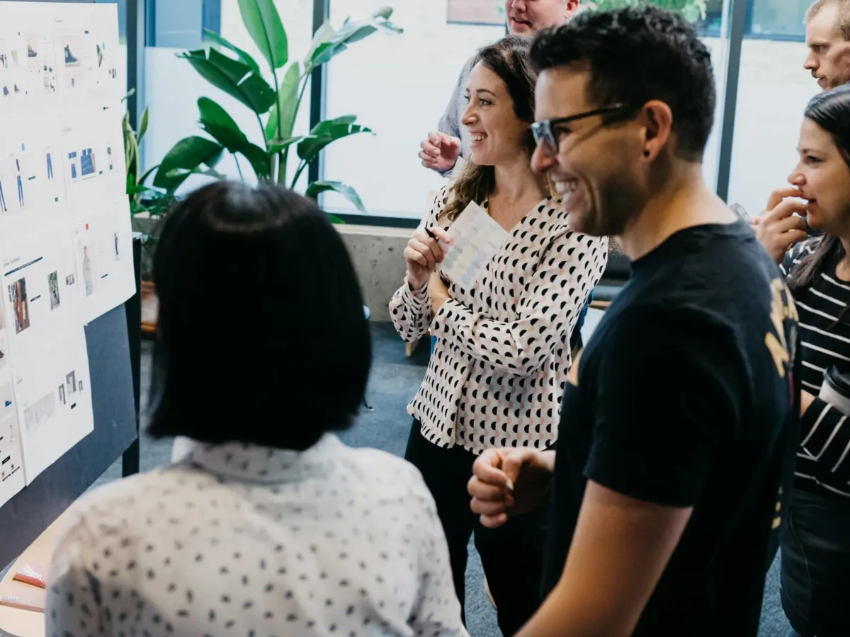 A group of people smiling and discussing ideas posted on a board in a bright office space.