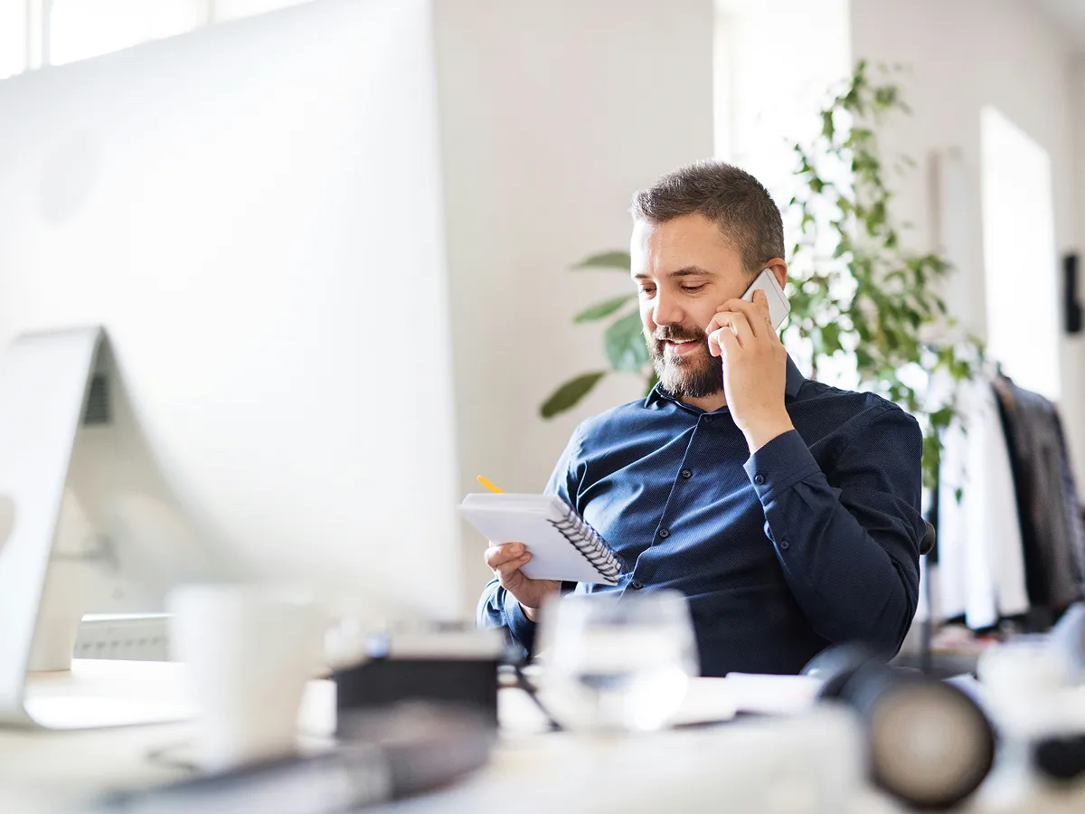 Bearded man in blue shirt talking on phone while writing notes in a bright office.