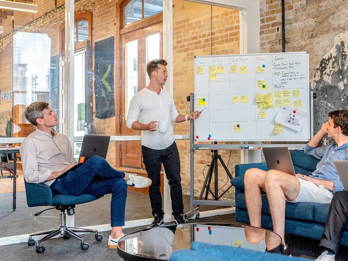 Man in blue shirt talking on phone while writing notes in a notepad at a bright office desk.