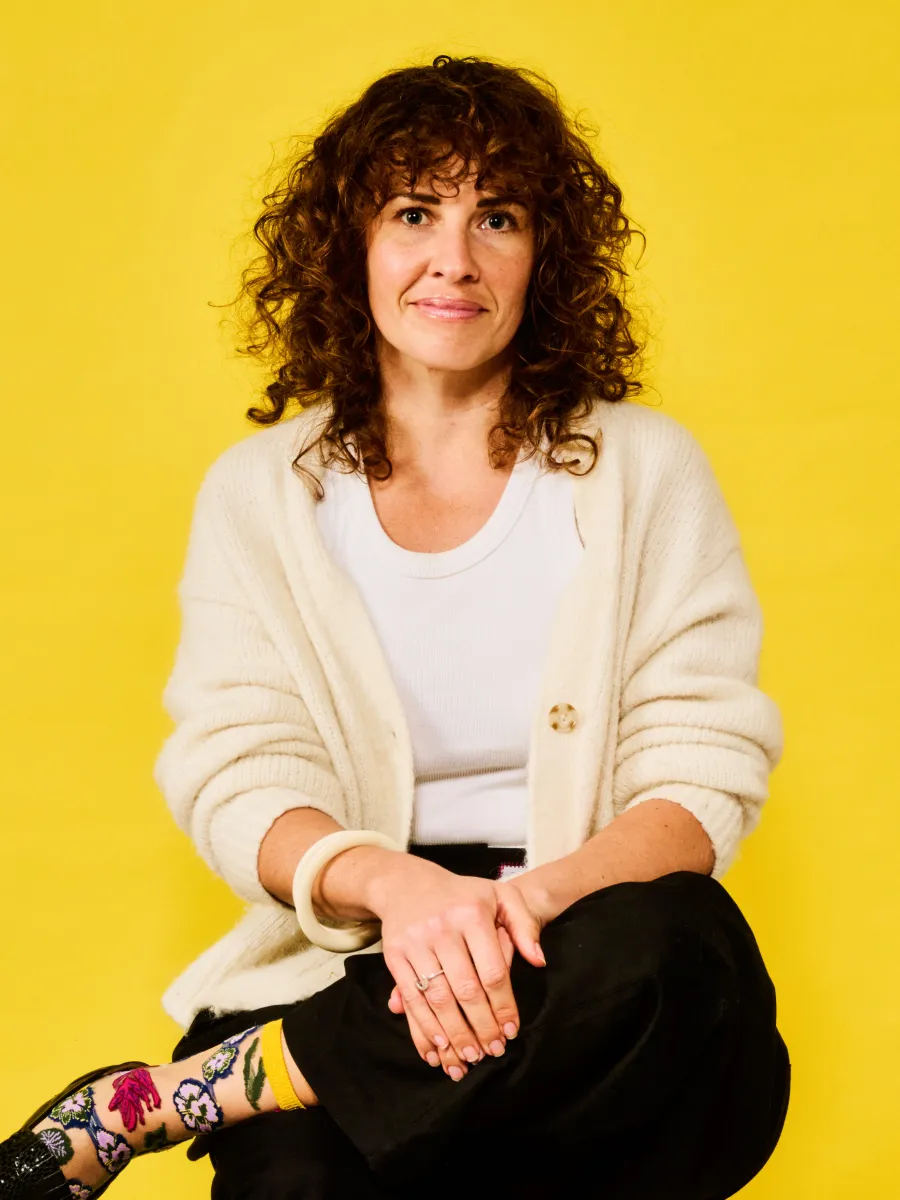 Woman with curly hair in white cardigan, black pants, and floral socks sitting against yellow background.