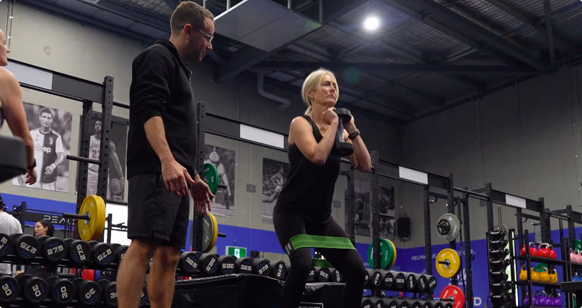 Trainer assists woman doing squats with weights and resistance band in a gym filled with equipment.