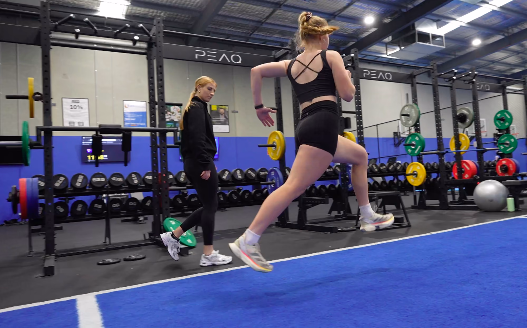 Athlete in black sportswear jumping indoors while a trainer watches in a gym with weight racks.