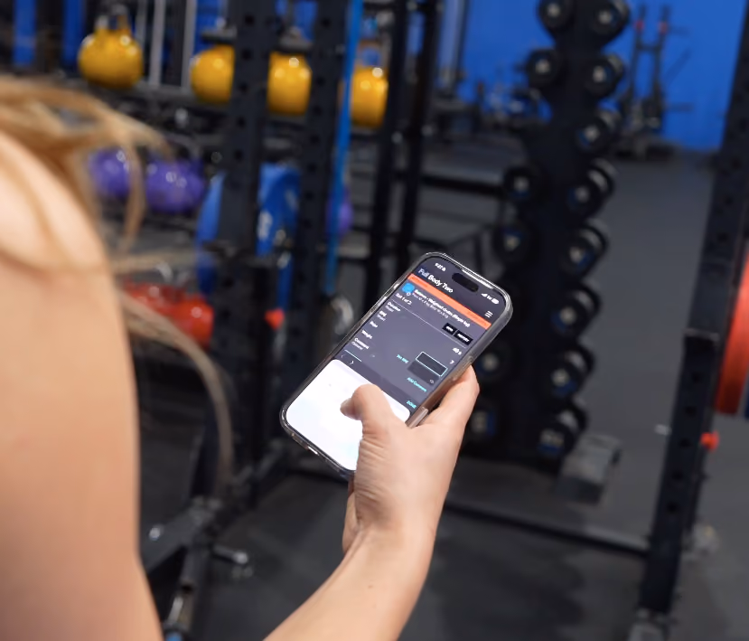 Person holding phone showing a workout app screen inside a gym with weights in the background.