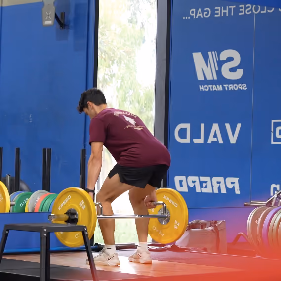 Athlete in maroon shirt and black shorts lifting a barbell with yellow weights in a gym.