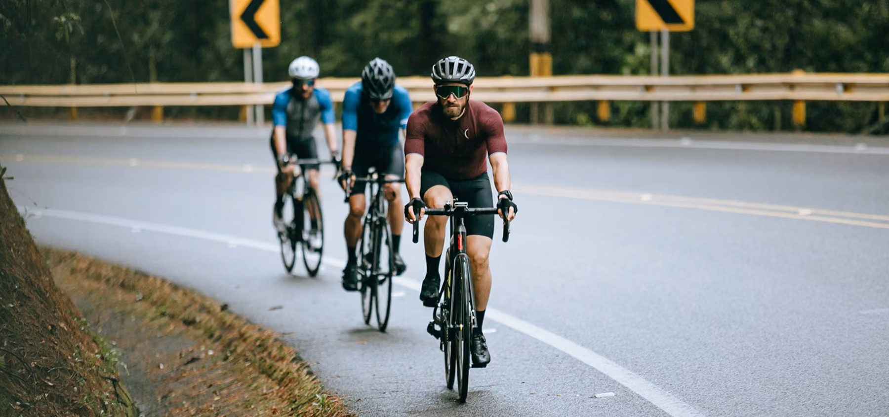 Three men in cycling gear riding road bikes on a curving, wet road with caution signs.