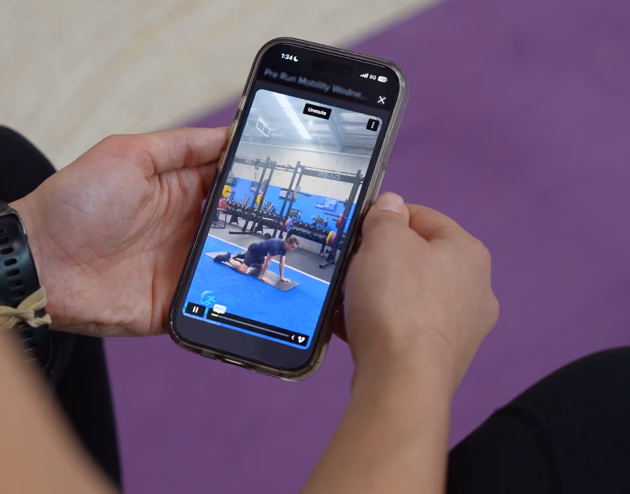 Person holding phone showing exercise video of a man doing stretches on a mat in a gym.