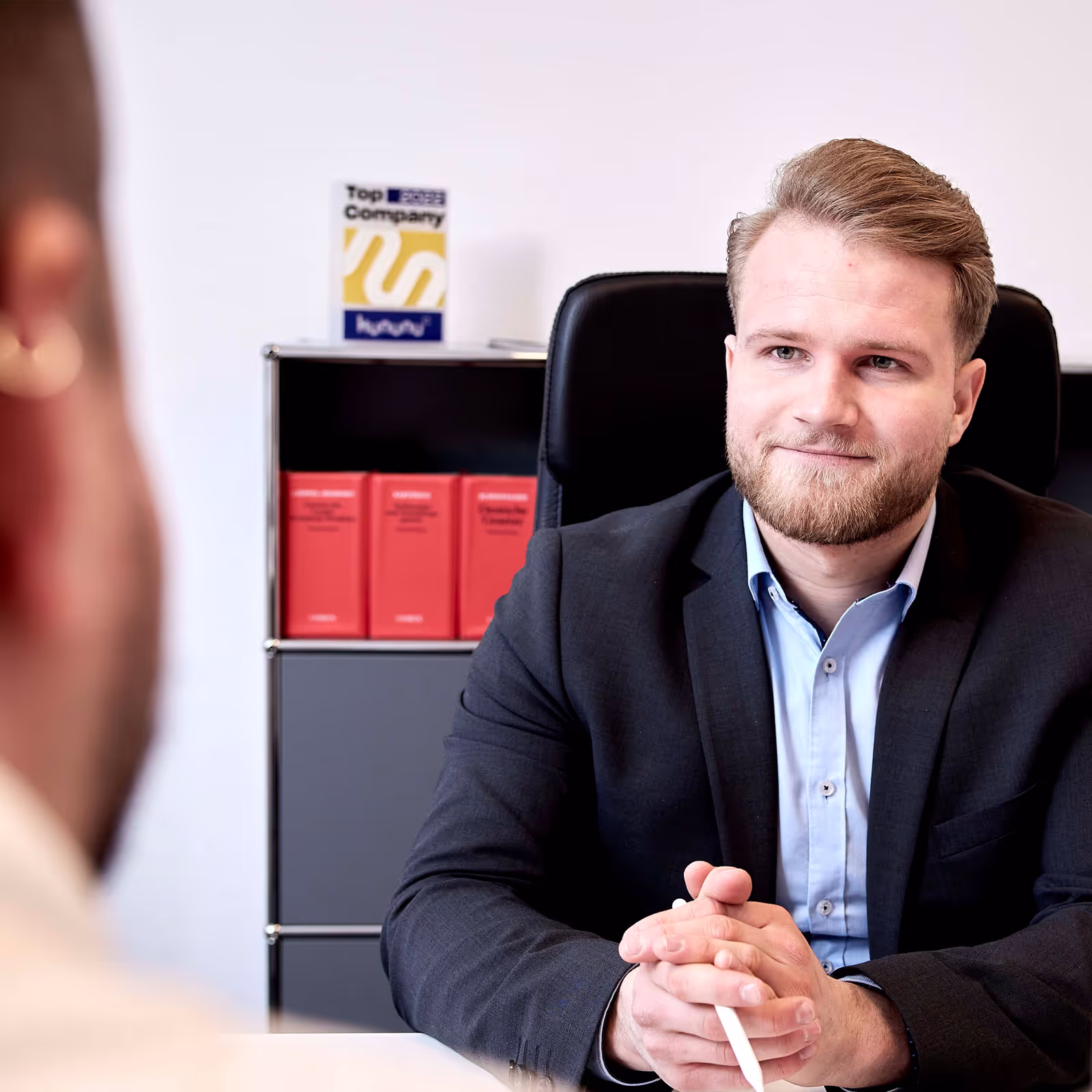 Ein professioneller Gesprächspartner im Business-Outfit führt ein Vorstellungsgespräch in einem modernen Büro mit Auszeichnung „Top Company 2023“ im Hintergrund.