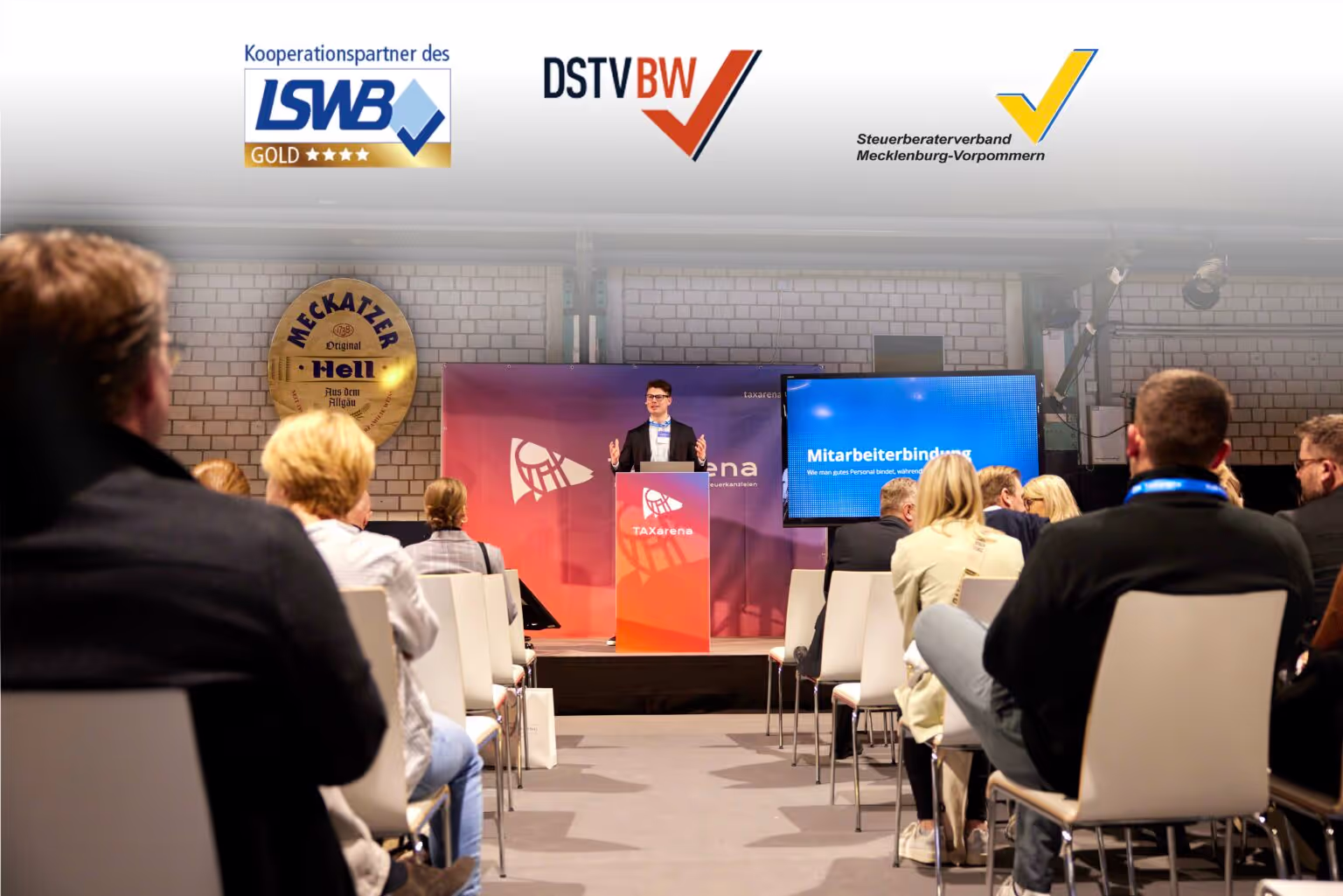 Ein Redner steht auf der Bühne der TAXarena vor einem Publikum und spricht über das Thema Mitarbeiterbindung, während Logos von Steuerberaterverbänden eingeblendet sind.