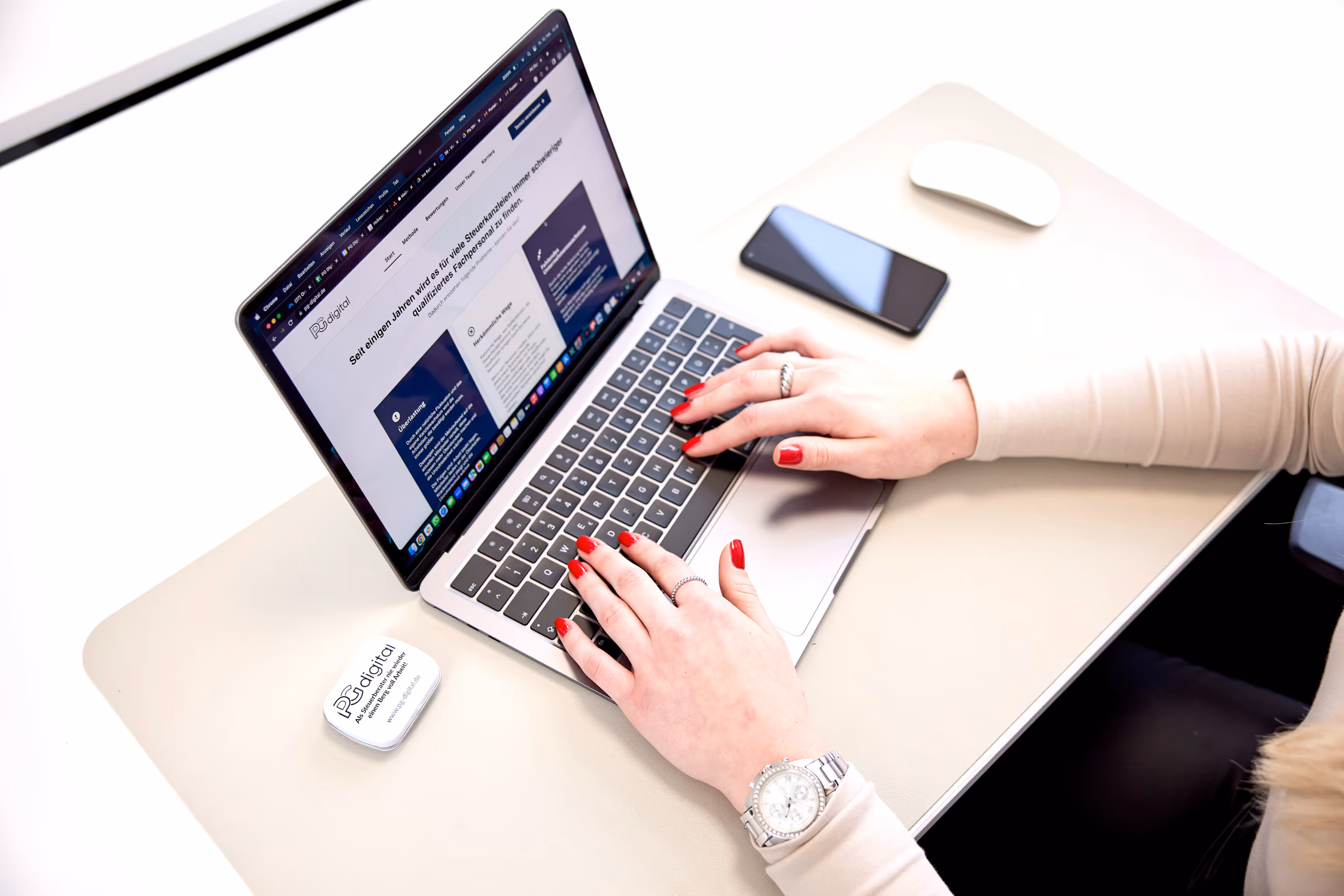 Nahaufnahme von Händen mit rotem Nagellack, die auf einem Laptop die Website von PGdigital bedienen. Auf dem Schreibtisch liegen ein Smartphone, eine Maus und ein PGdigital-Giveaway.