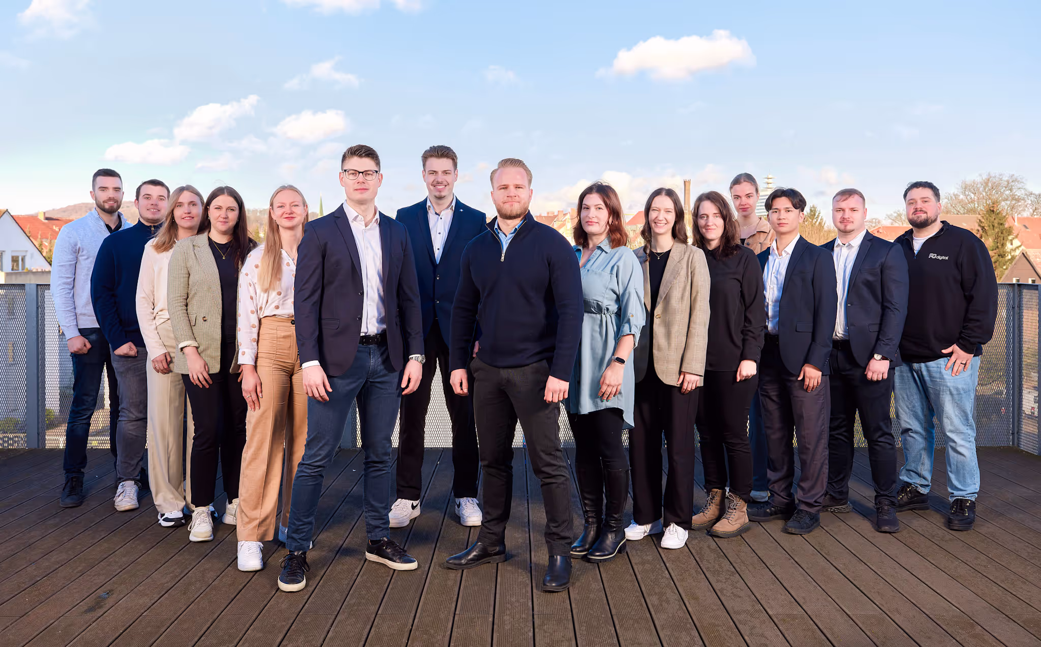 Gruppenfoto eines jungen Teams von PGdigital auf einer Dachterrasse bei sonnigem Wetter. Die Personen stehen nebeneinander vor einem Geländer mit Blick auf Dächer und blauen Himmel im Hintergrund.