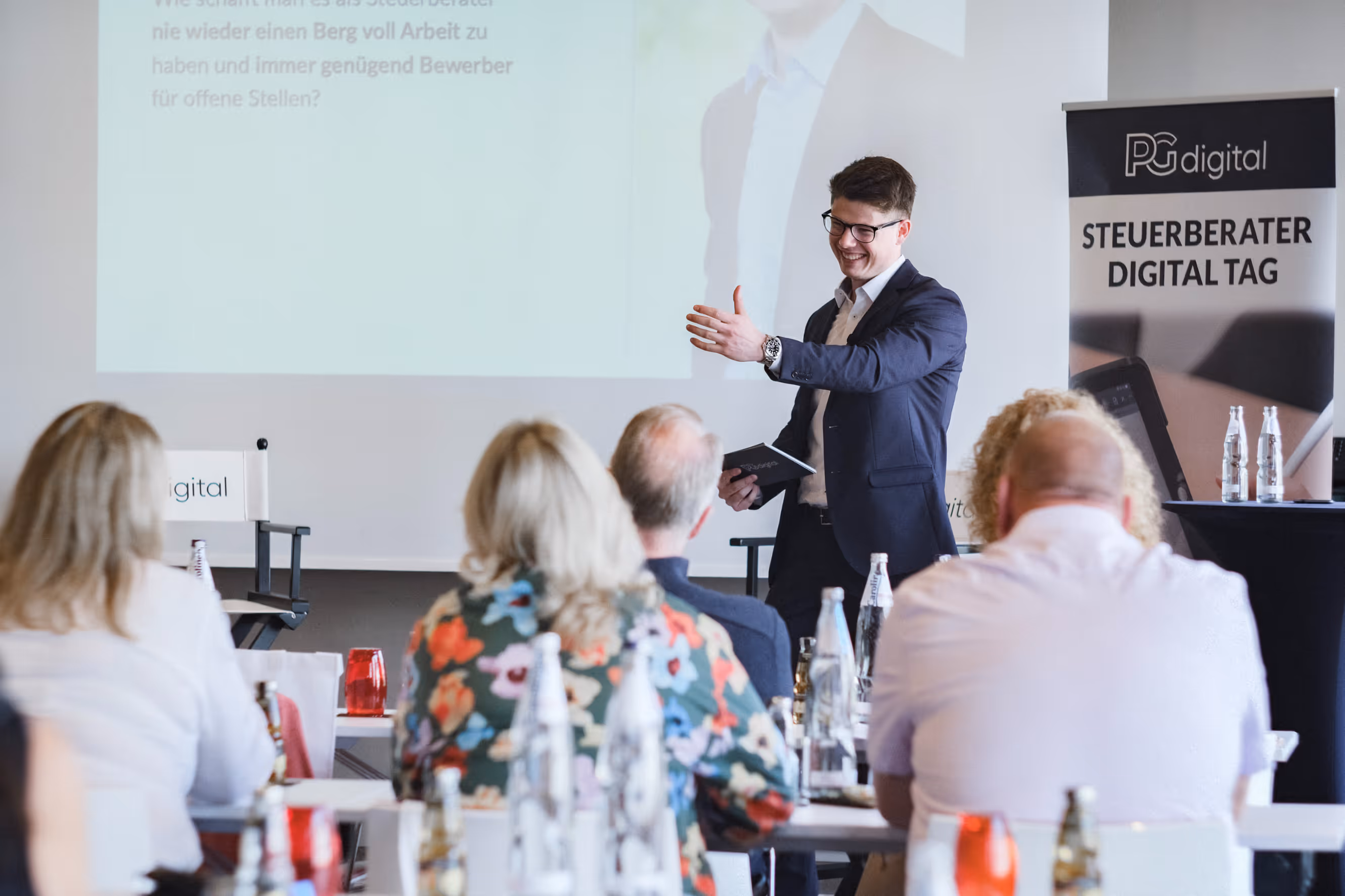 Ein Redner hält eine Präsentation auf dem Steuerberater Digital Tag vor einem sitzenden Publikum. Im Hintergrund ist eine Leinwand mit Projektion und ein Banner mit der Aufschrift „PG digital – Steuerberater Digital Tag“ zu sehen.