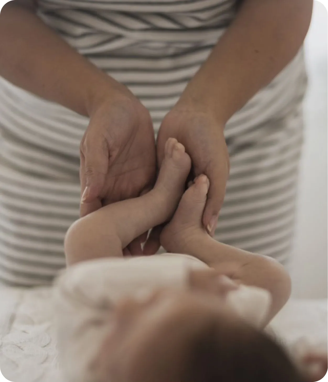 Un adulte tenant doucement les pieds d’un bébé allongé, avec un fond de vêtements rayés.