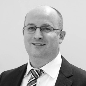 Smiling bald man wearing glasses, a suit, and a striped tie on a plain background.