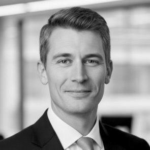 Headshot of a man with short hair wearing a suit and tie, smiling slightly with blurred background.