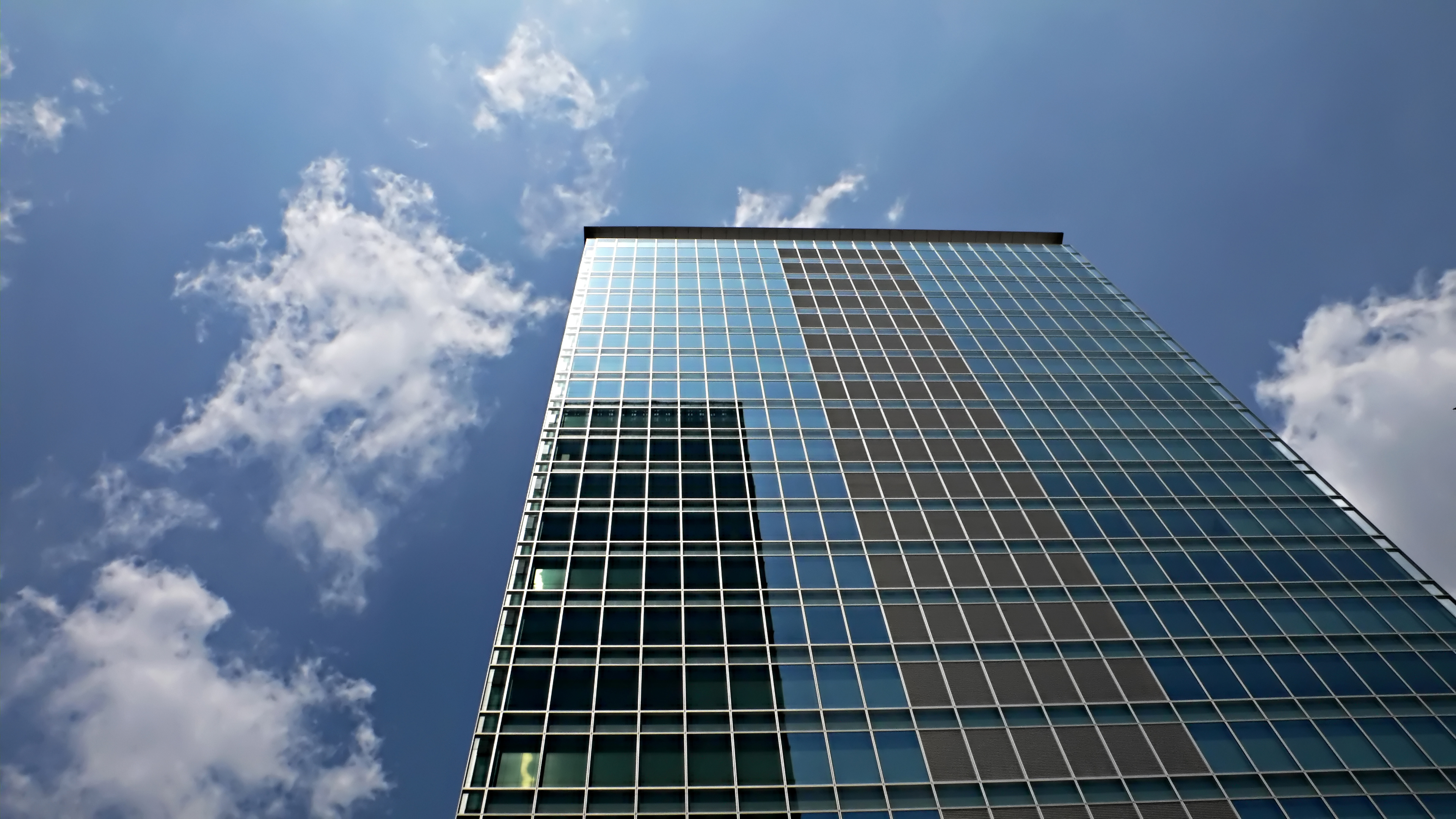 Looking up at a tall modern glass building reflecting blue sky and clouds.