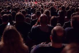 Large audience seated closely together, facing forward, with many people visible from behind in a dimly lit setting.