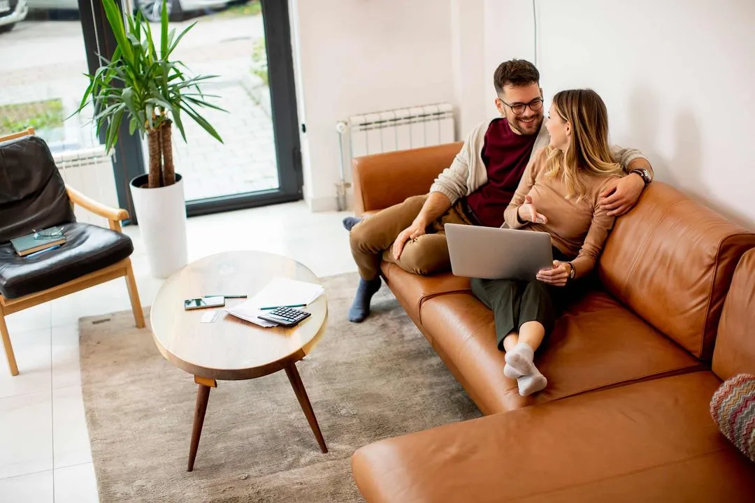 A couple sits on a couch, using a laptop together and smiling.