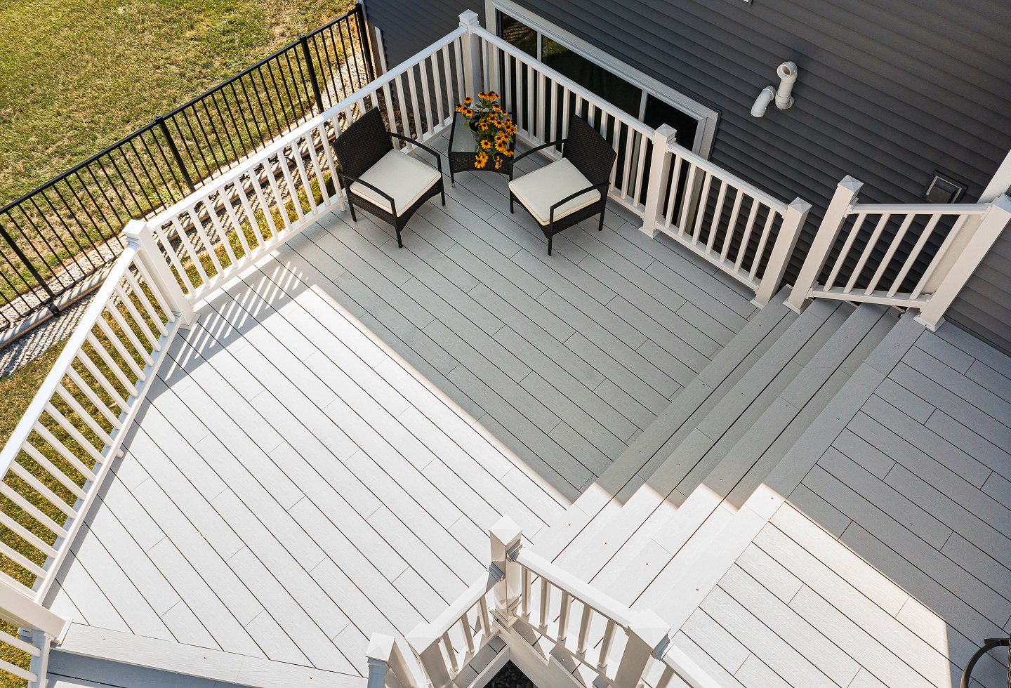 White and gray wooden porch deck with two wicker chairs and a small table with yellow flowers.