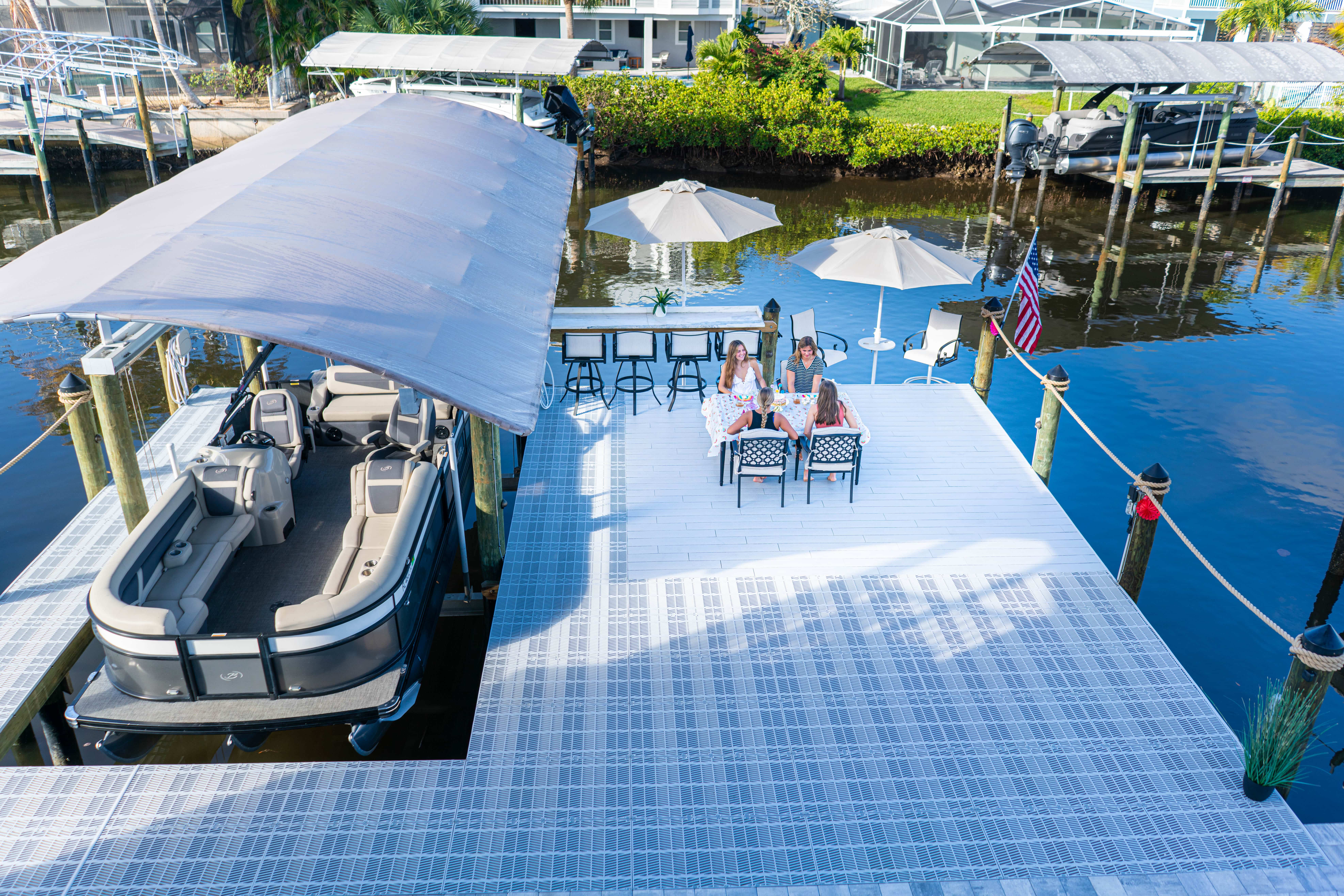 Four people sitting at table with umbrellas on dock next to covered boat on calm water.