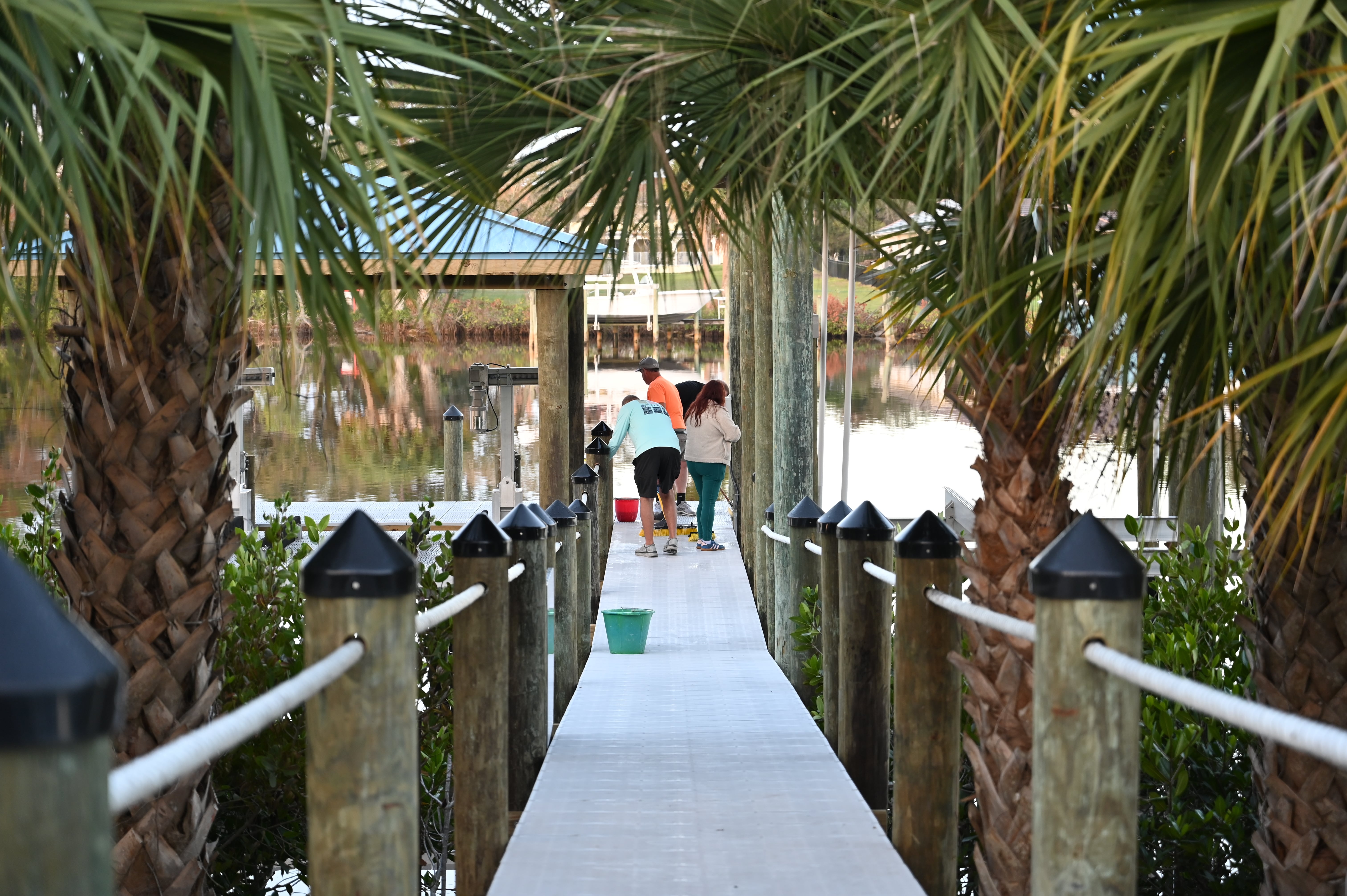 Three people standing on a dock under palm fronds by calm water with boats in the background.