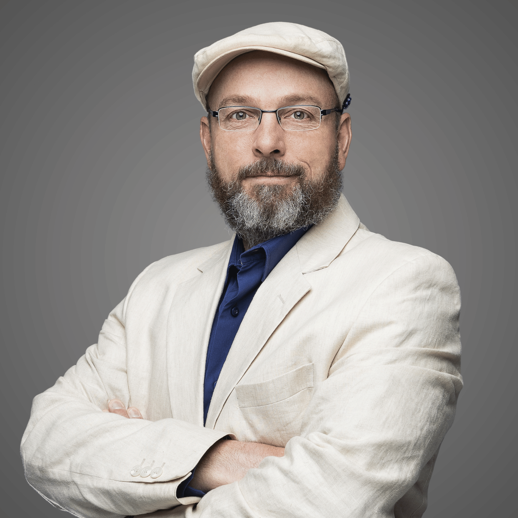 Man with beard and glasses wearing a beige cap and jacket over a navy shirt with arms crossed.