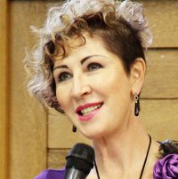 Smiling woman with curly short hair speaking into a microphone in front of wooden background.