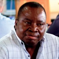 Man in a white checkered shirt sitting indoors with a thoughtful expression.