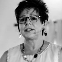 Woman with short hair, glasses, speaking, wearing earrings and a necklace, black and white photo.