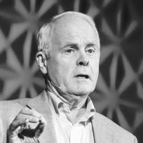 Older man in a light jacket speaking with a serious expression against a geometric background.