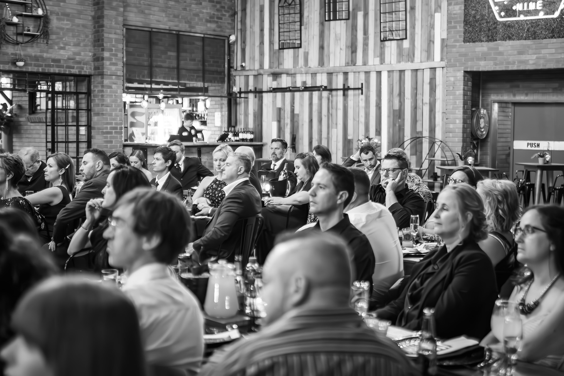 Audience of men and women in formal attire seated and attentively listening indoors at an event.