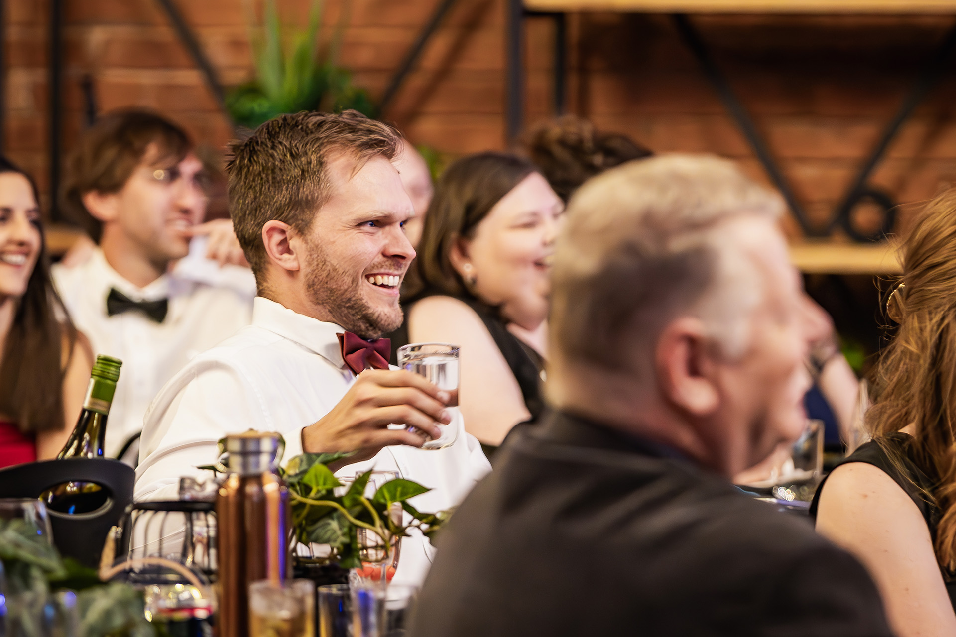 Man in white shirt and red bow tie laughing and holding a clear glass at a social gathering.