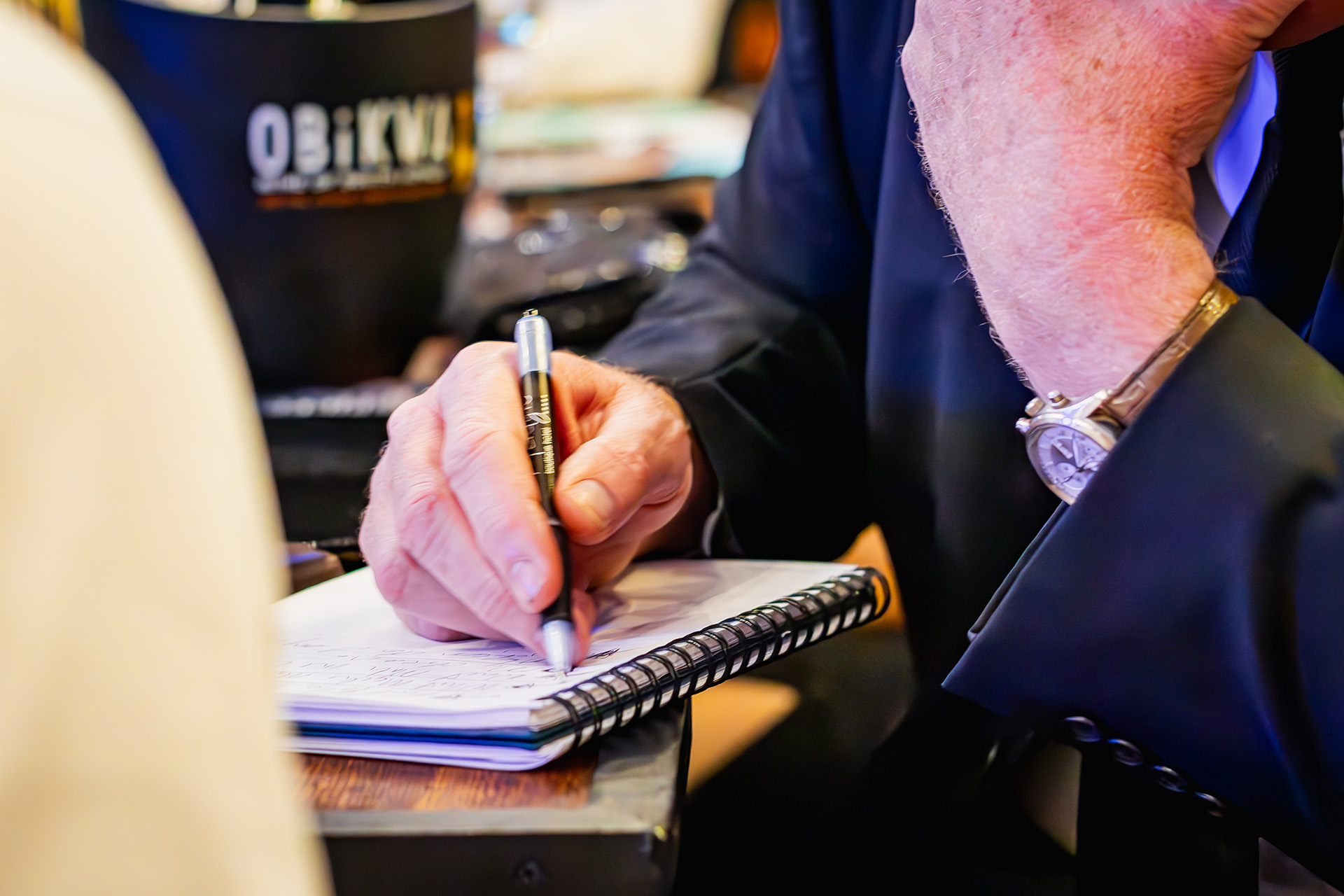 Close-up of an elderly man's hand writing with a pen in a spiral notebook.