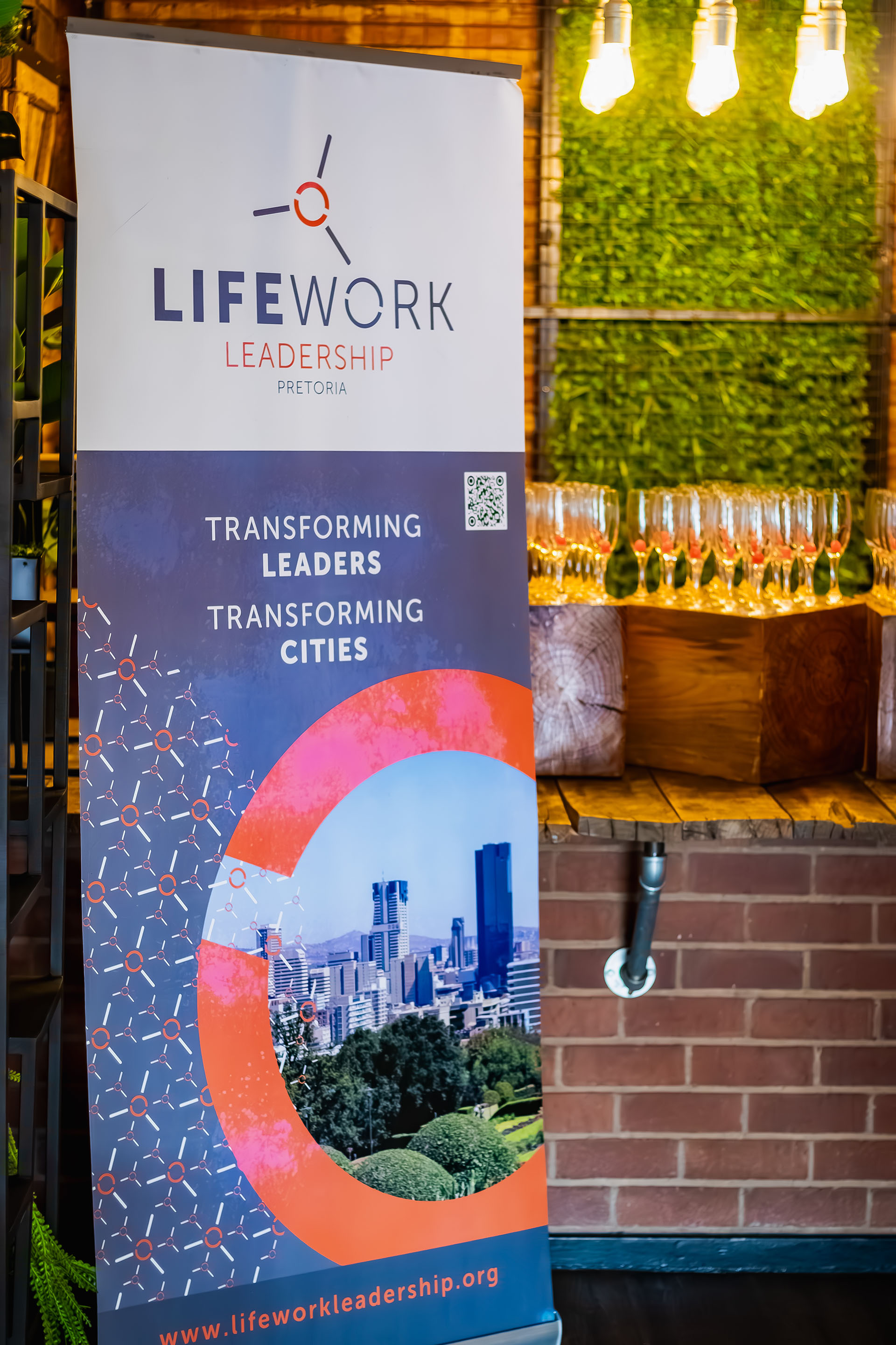 LifeWork Leadership banner with city skyline, slogan, and website URL in indoor setting with glasses and lights.