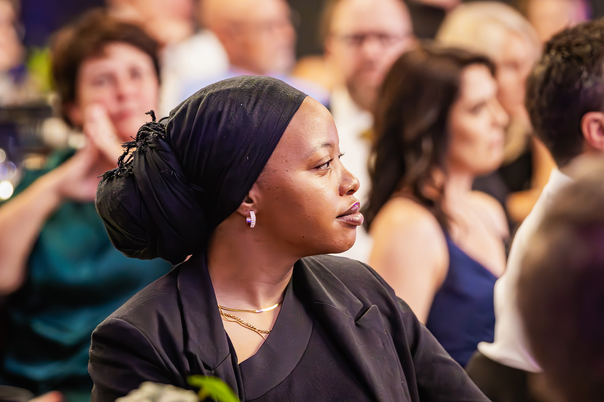 Woman in a black headscarf and dark clothes attentively listening at a formal event.