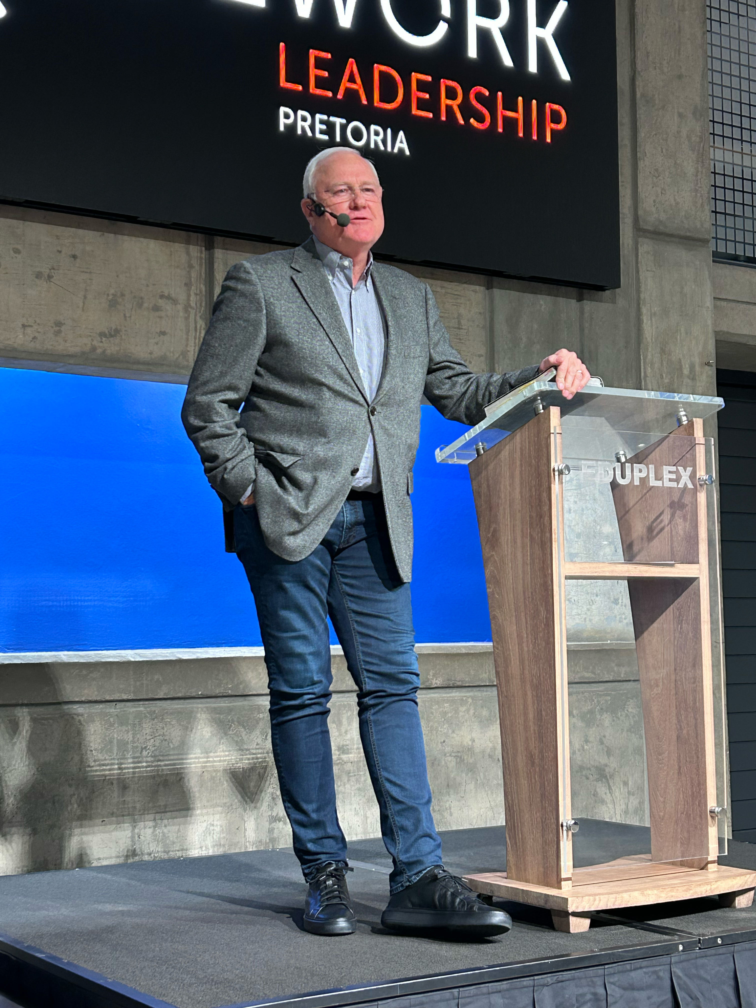 Man in gray blazer and jeans speaking on stage with a headset mic, standing by a wooden podium.