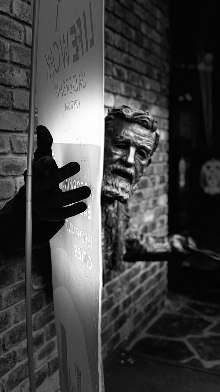 Black and white photo of a sculpture of a man peeking around a sign attached to a brick wall.
