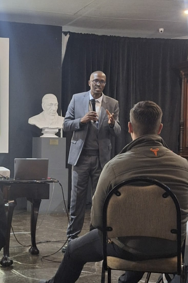 Man in a gray suit speaking with a microphone to a seated audience member in a room with a bust statue.