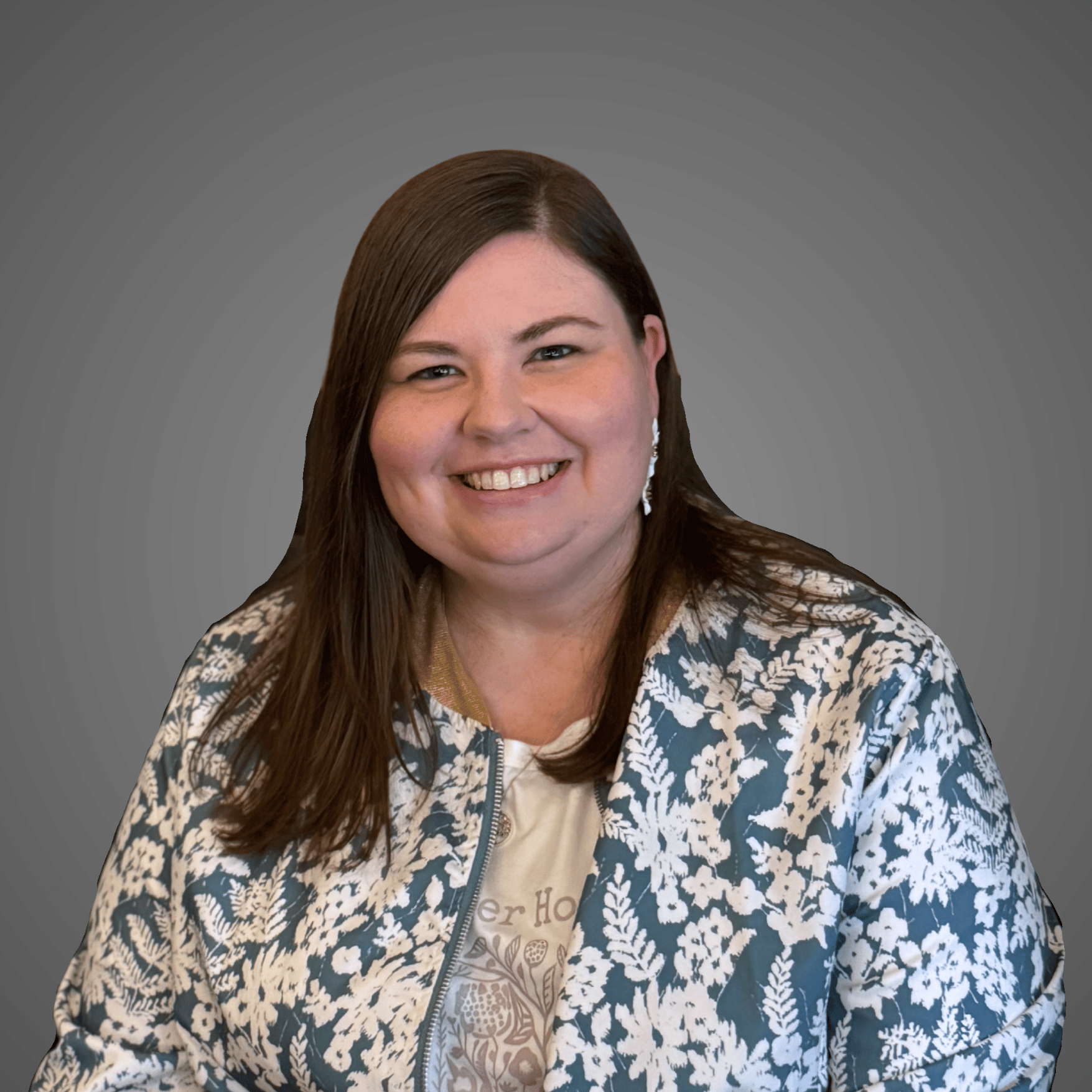 Smiling woman with long brown hair wearing floral patterned jacket and white earrings on gray background.