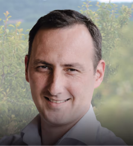 Man with short dark hair smiling outside with greenery and hills blurred in the background.