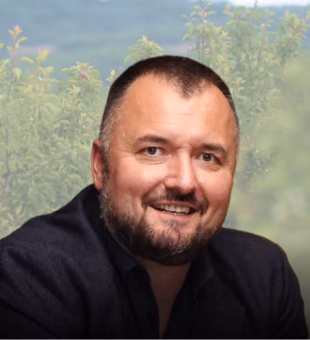 Smiling man with short dark hair and beard wearing a black shirt with greenery in the background.