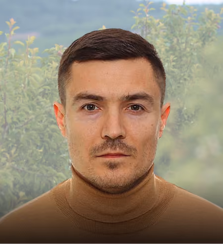 Man with short dark hair and brown turtleneck sweater standing outdoors with blurred green foliage background.