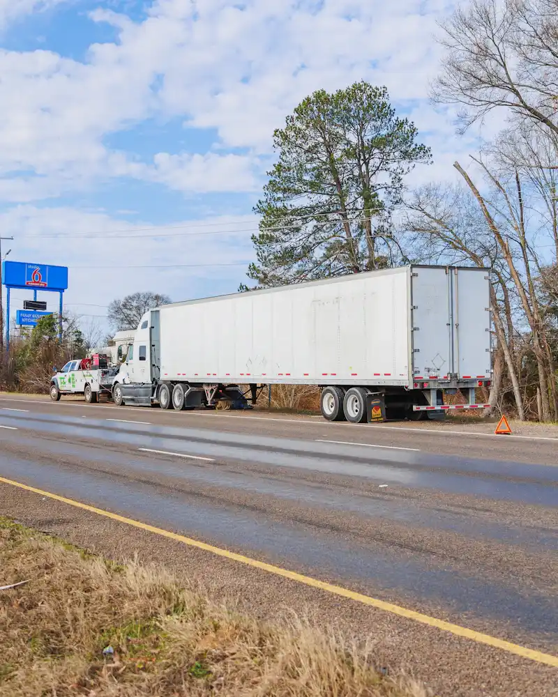 Roadside service assisting a disabled semi-truck on the highway shoulder with reflective warning triangle deployed. A Studio 6 sign is visible nearby.