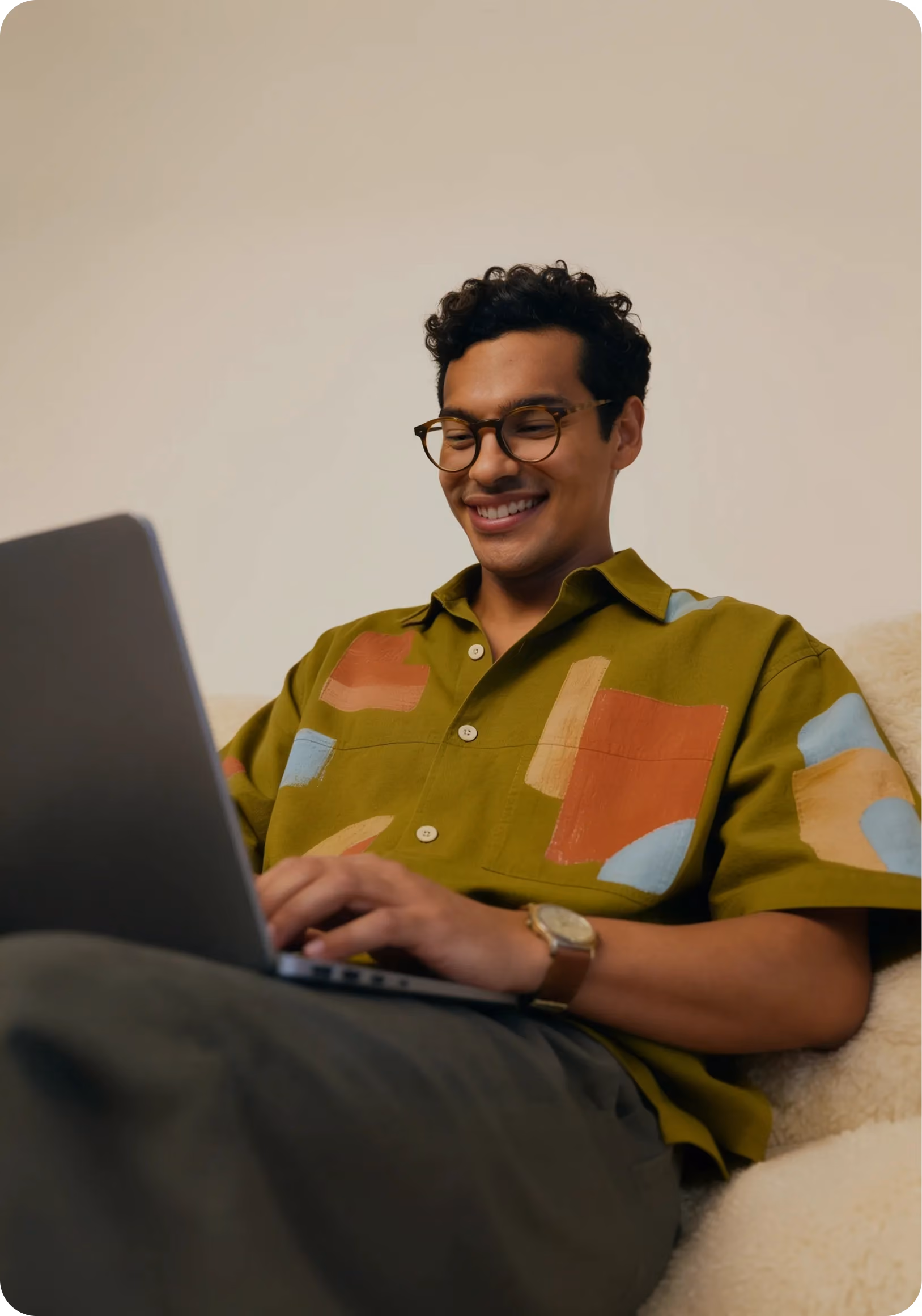 Smiling man with glasses using a laptop while sitting comfortably on a beige couch.