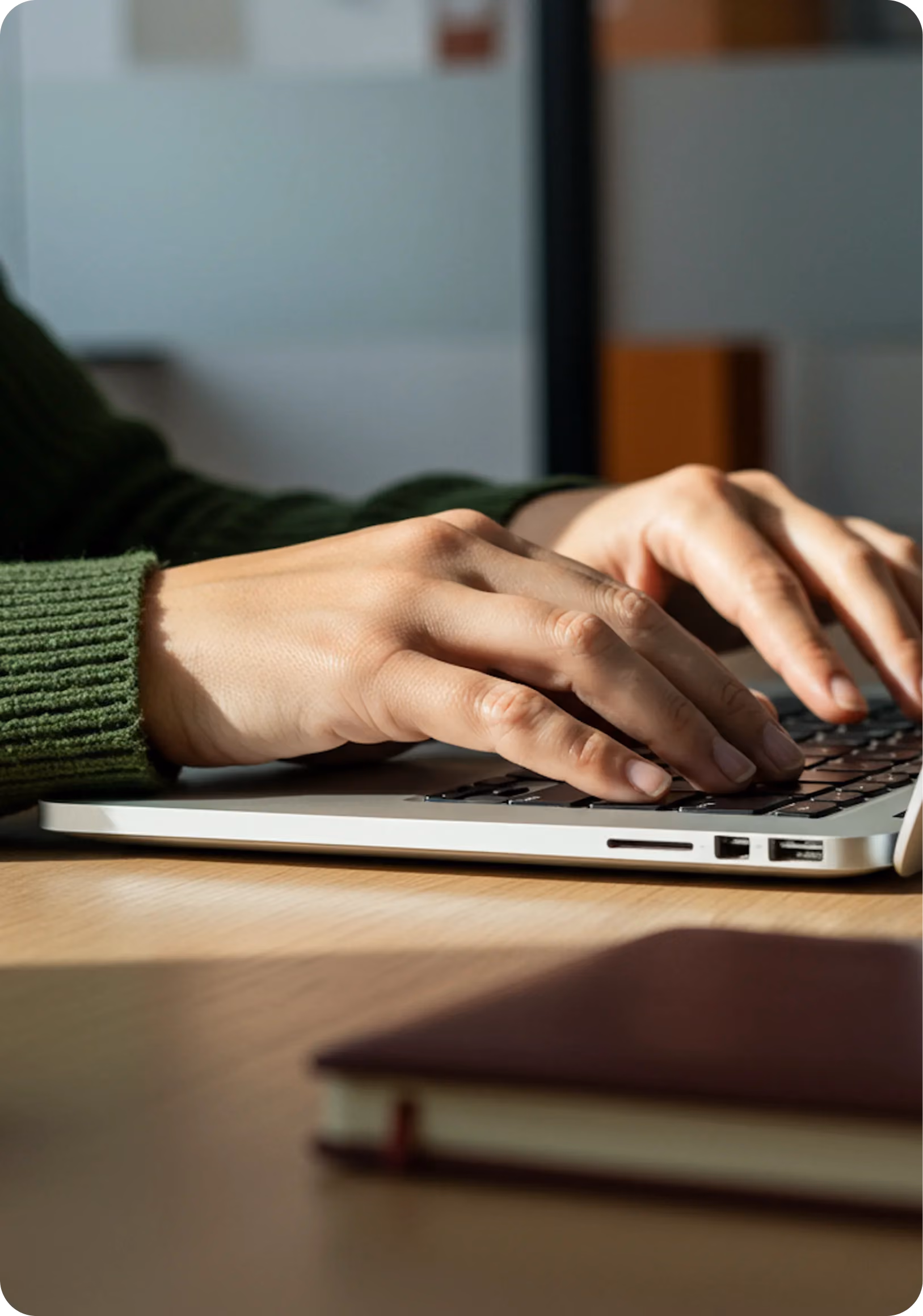 Hands typing on a laptop keyboard with a notebook blurred in the foreground on a wooden desk.