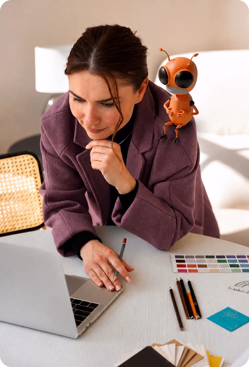Woman in purple jacket working on laptop with color swatches and pencils on table, cartoon bug on shoulder.