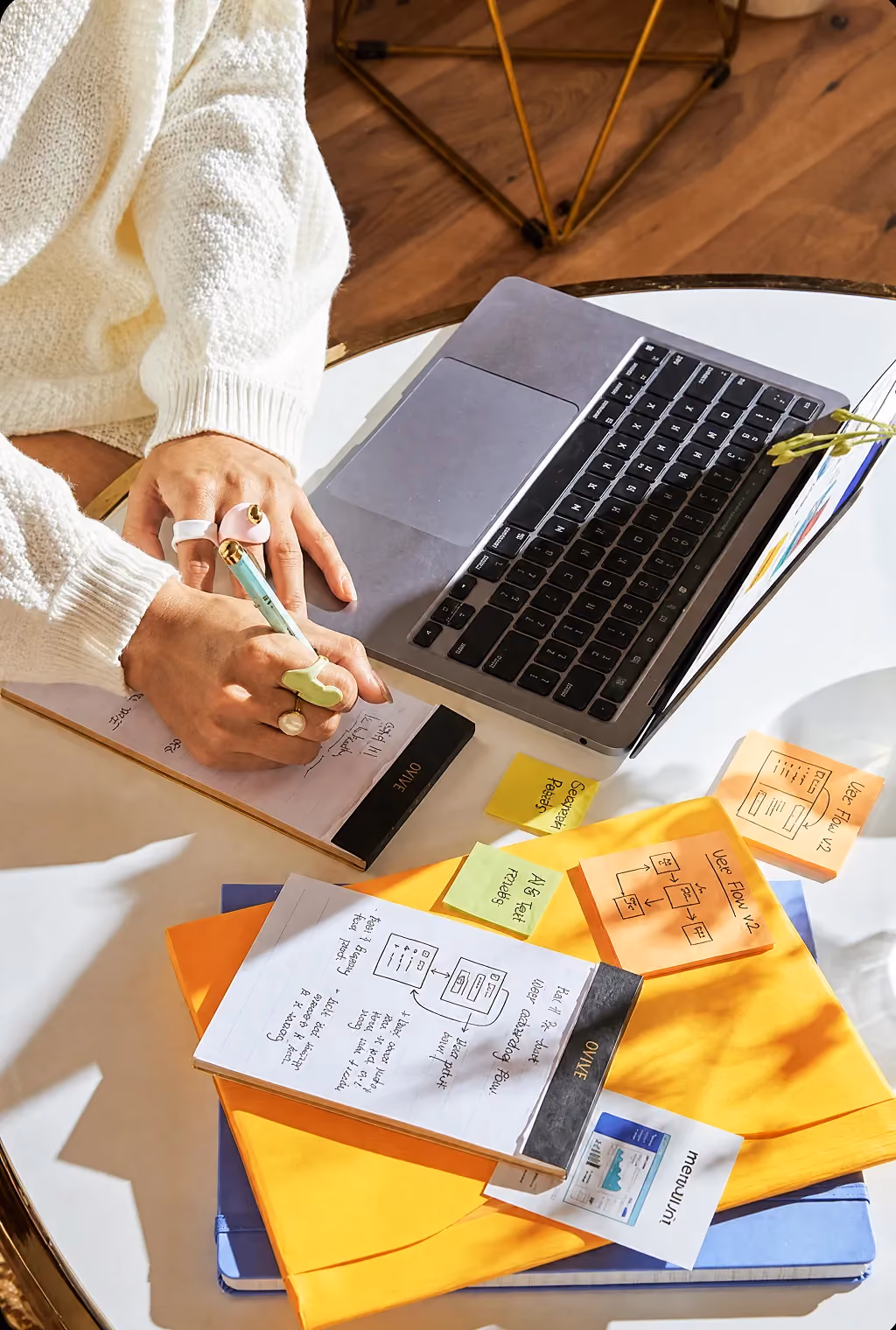 Person in white sweater writing notes beside a laptop and colorful sticky notes on a glass table.